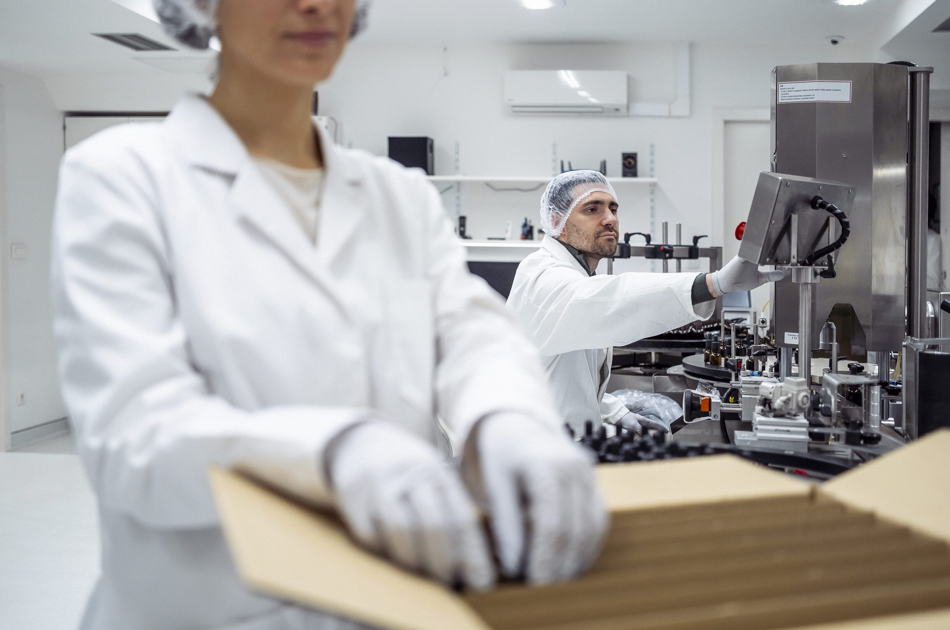 A mid adult Caucasian pharmacy worker in a modern laboratory operates an advanced extraction system. Wearing a white lab coat, gloves, and a hair net, the worker displays focus and precision in pharmaceutical procedures.